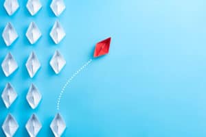 Featured image of blogpost "Finding What Makes You Happy - A Personal Account" with a light blue background, white paper boats and a red paper boat changing path from comformity