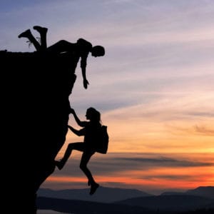featured image of "The Art of Guidance: Coaching vs. Mentoring" showing a cliff, somebody reaching down to help another person up and a sunset in the background