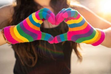 Featured image of blogpost "The Importance of Pronouns" showing two hands in rainbow-colored gloves making the heart symbol.